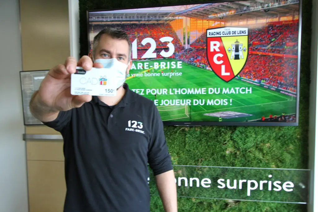 123PareBrise-Lens-joffrey-carte-cadeau Chef de centre Joffrey qui tient une carte cadeau de 150€ dans sa main devant un écran RC Lens