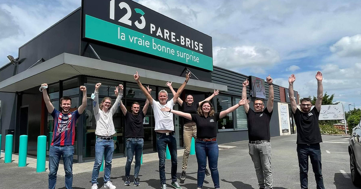 Denis Brogniart et les collaborateurs 123 Pare-Brise prennent une photo fun ensemble devant l'agence 123 Pare-Brise Arras.